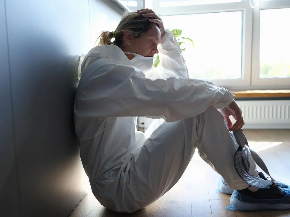 Tired female doctor in medical protective suit and mask in hands after work in clinic. Harsh working conditions of doctors in coronavirus pandemic concept