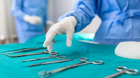 Nurse hand taking surgical instrument for group of surgeons at background operating patient in surgical theatre. Steel medical instruments ready to be used. Surgery and emergency concept