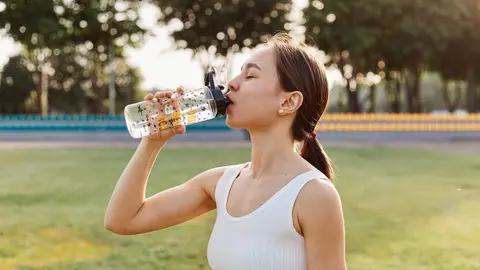 Fit dark haired female drinking water from her water bottle on stadium just before she starts training, outdoor workout in summertime, healthy lifestyle.